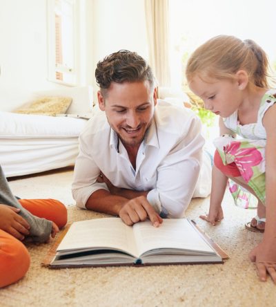 Family reading a book on the floor in the living room. Father reading stories to his son and daughter at home. Young man, little boy and little girl reading story book.
